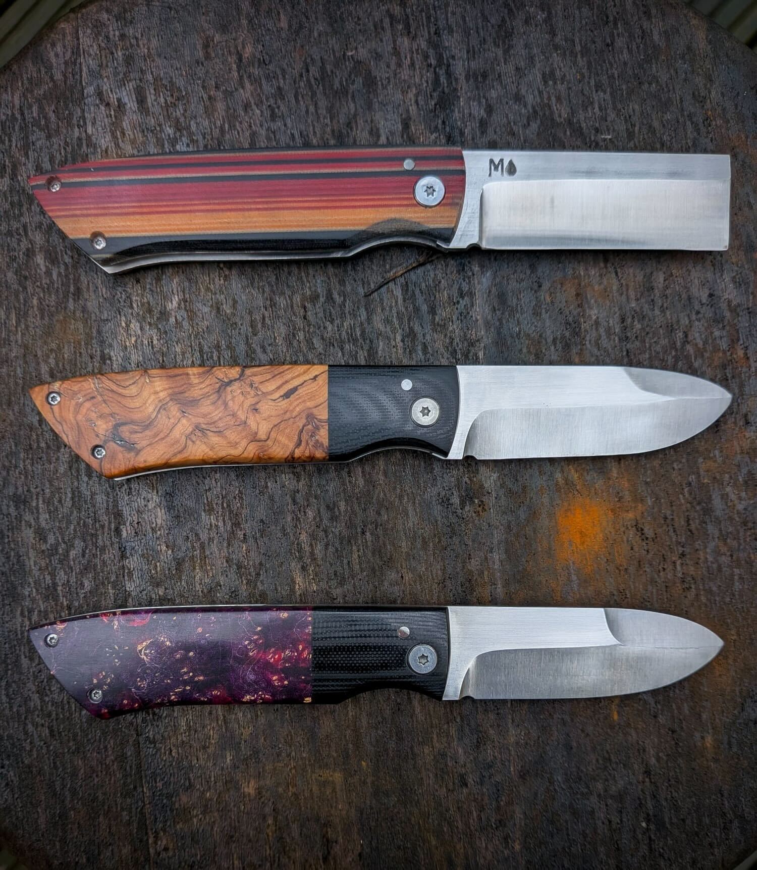 Three handmade folding knives with blades open, featuring striped wood, olive wood, and resin handles